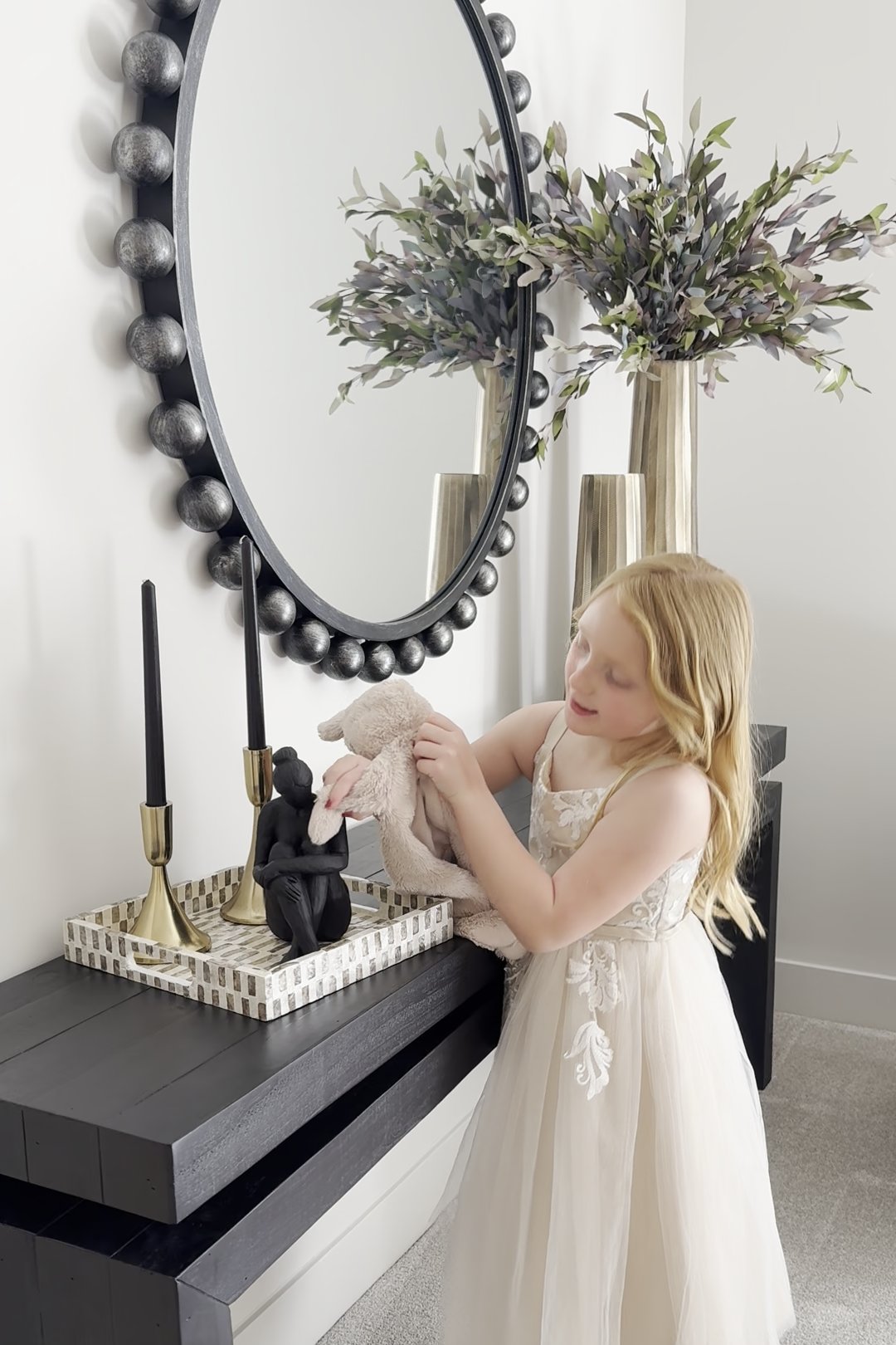 A young girl in a tulle dress holds her stuffed bunny beside a black console styled with a beaded mirror, candlesticks, and olive branches