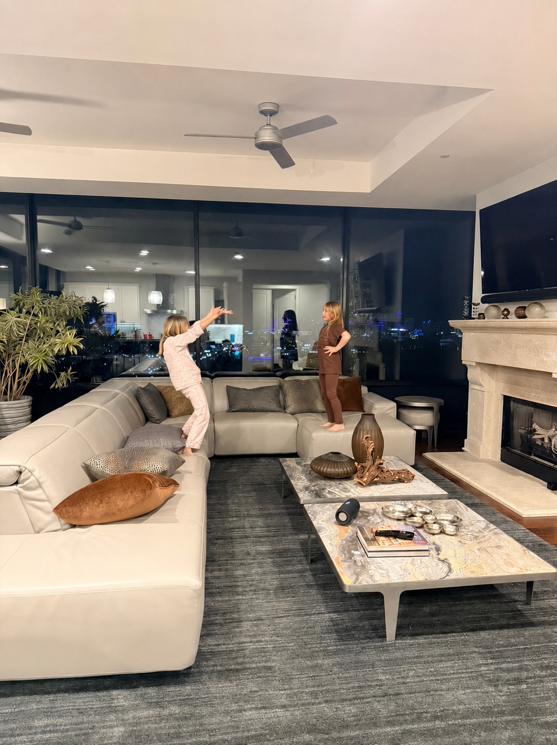 Two children play on a cream leather sectional in a warm, lamplit great room with marble cocktail tables and a city-lights window wall