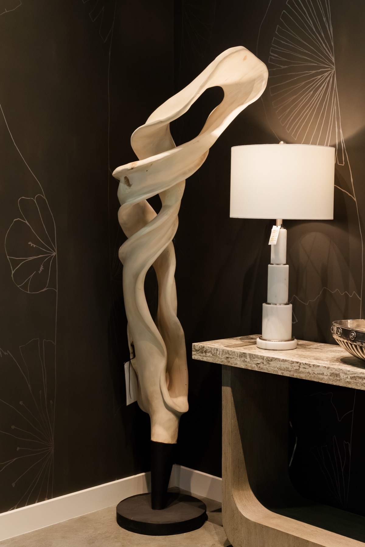 A sculptural carved-wood piece beside a stacked-marble lamp on a travertine console, against the hand-drawn ginkgo wall of the Bernhardt gallery.